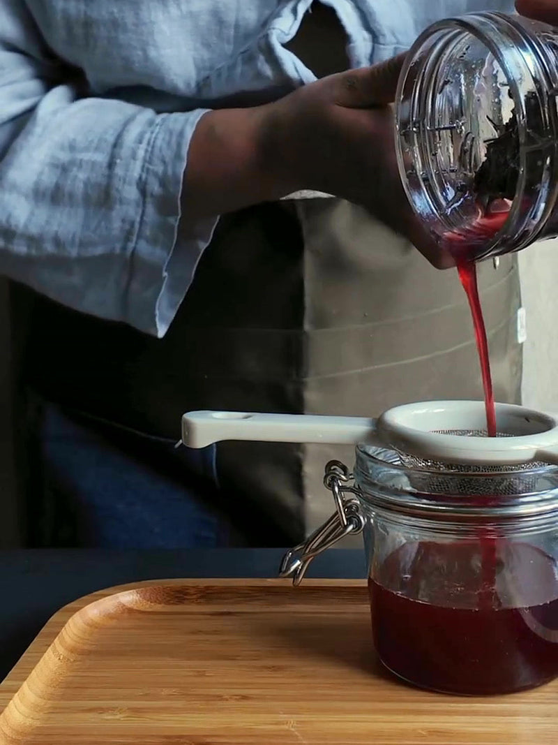 Oximiel rojo con flor de Jamaica pasando por un colador a un frasco de vidrio